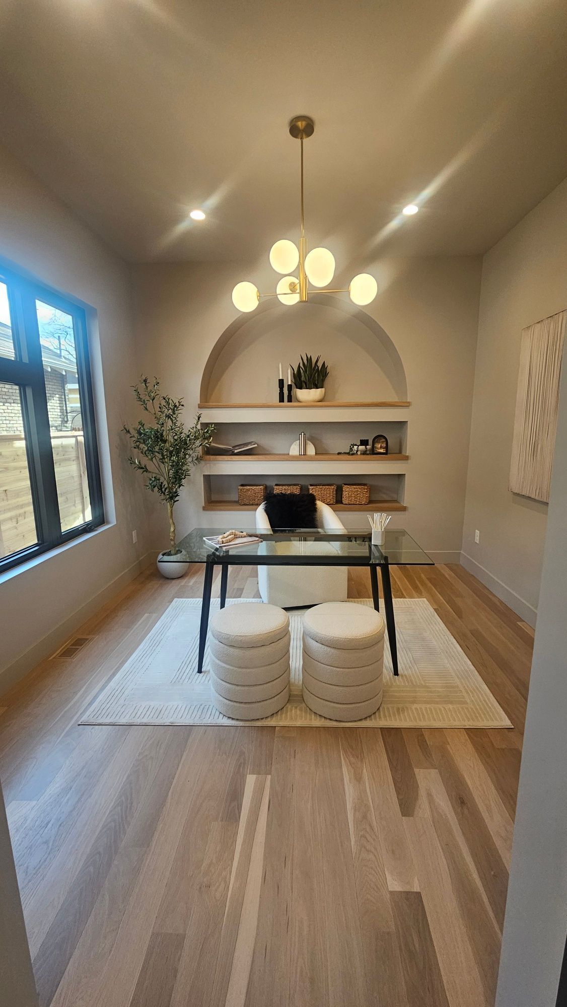A modern dining area with a glass table, two tufted stools, an arched built-in shelf, light wood floors, and a chandelier.