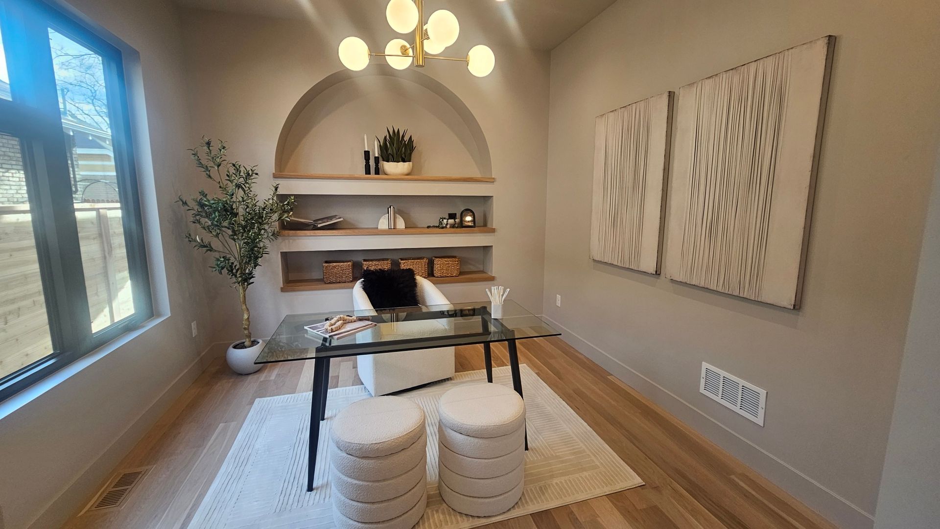 A modern, neutral-toned home office with an arched alcove, glass desk, two poufs, a potted plant, and wall art.