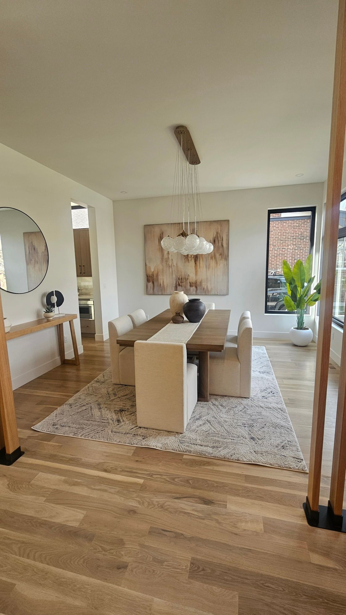 A dining room with a wooden table, light-colored chairs, a textured area rug, and hanging pendant lights.