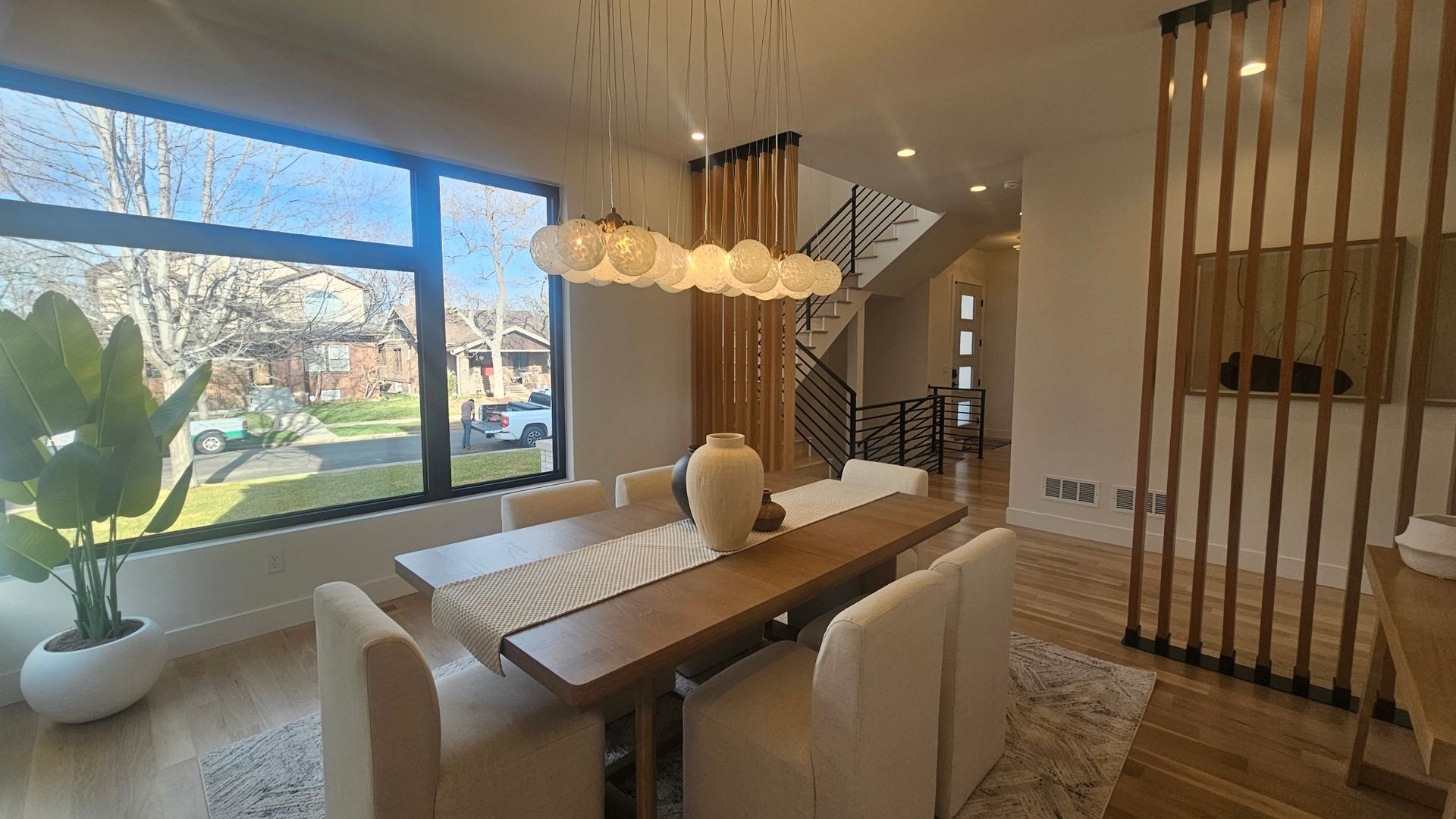 A modern dining room with a long wooden table, six light-colored chairs, a bubble chandelier, and tall wooden slat walls.