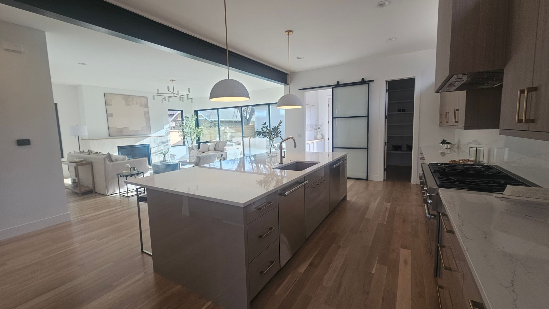 A modern kitchen with a large gray island, stainless steel appliances, and wood floors, opening into a bright living room.