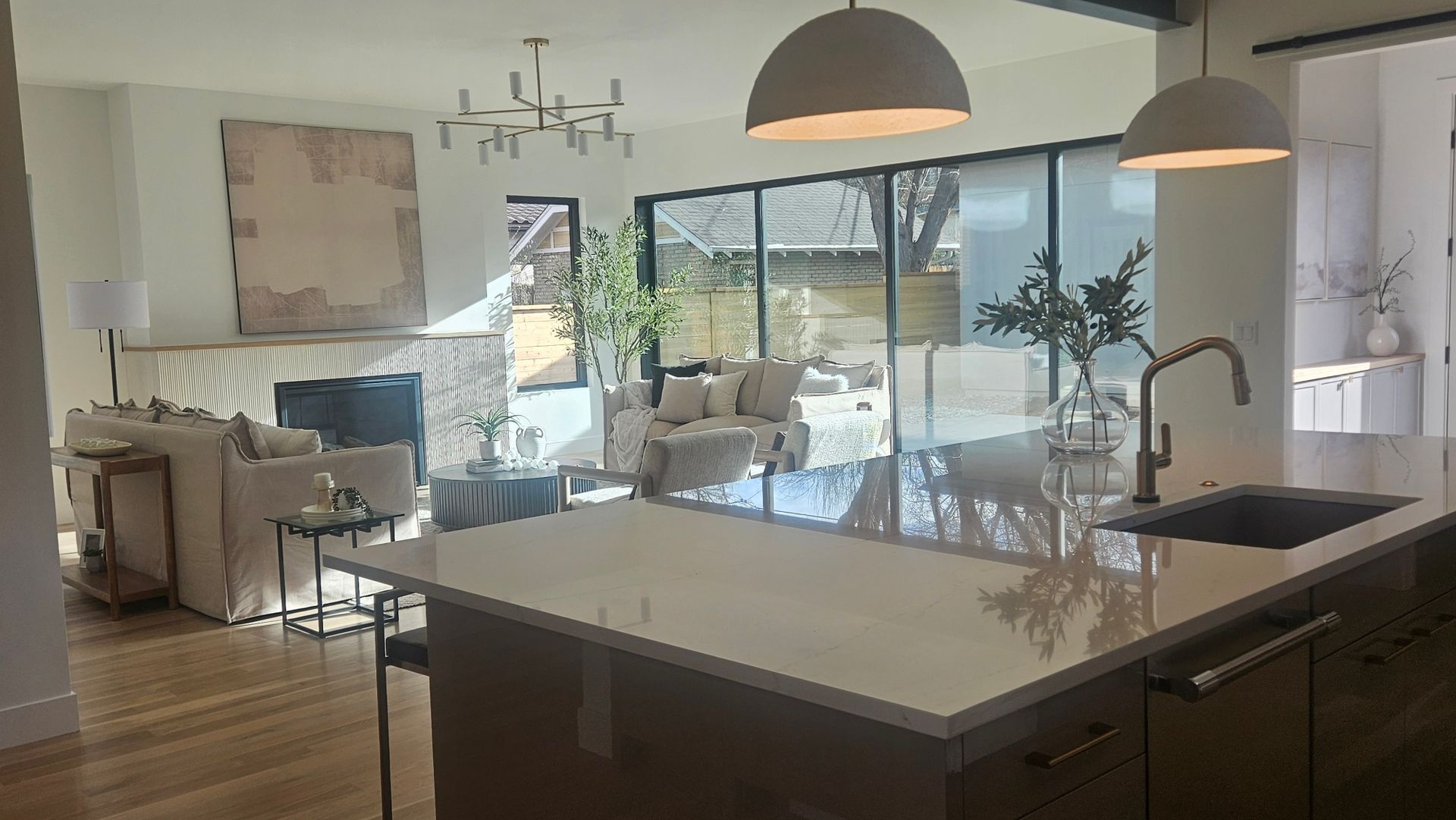 A modern, open-concept living space with a kitchen island in the foreground and a sunlit living room with large windows.