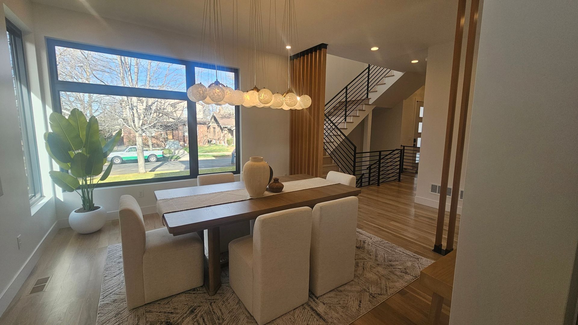 Dining area with a wooden table, six beige chairs, a pendant light fixture, and a large plant by a window.