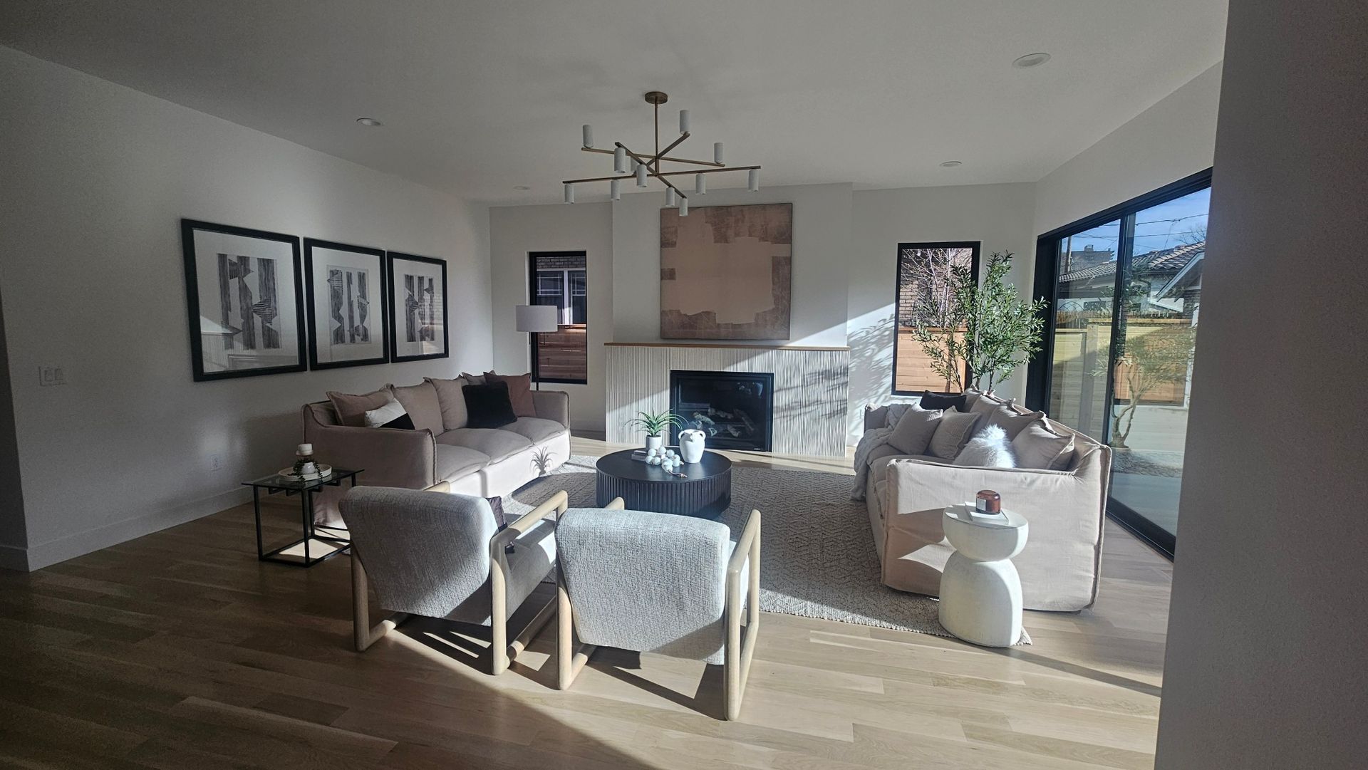A modern living room with two beige sofas, two textured armchairs, a central fireplace, and three framed wall prints.
