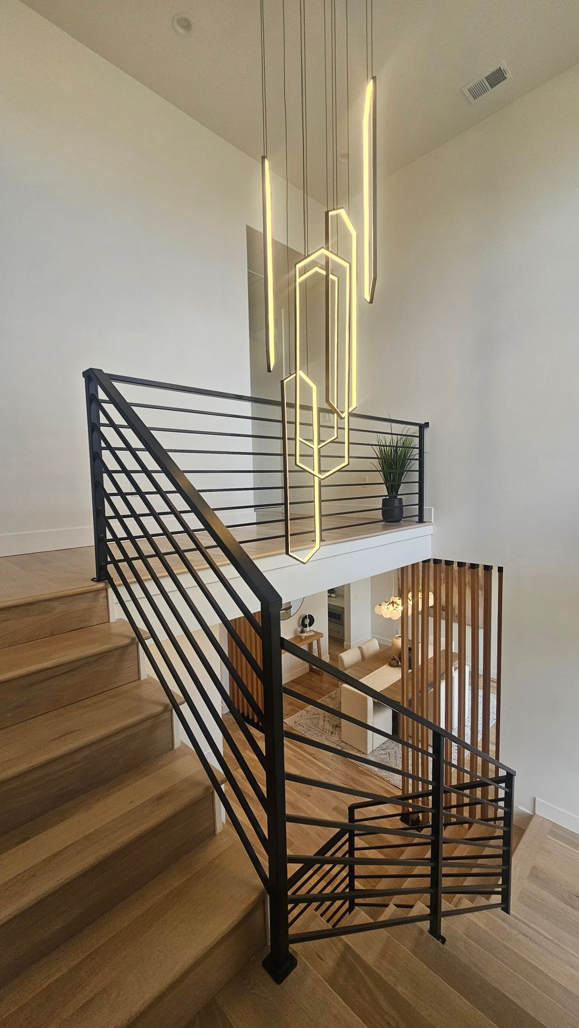 A modern staircase with black horizontal railings and light wood steps, featuring a tall, geometric LED chandelier above.