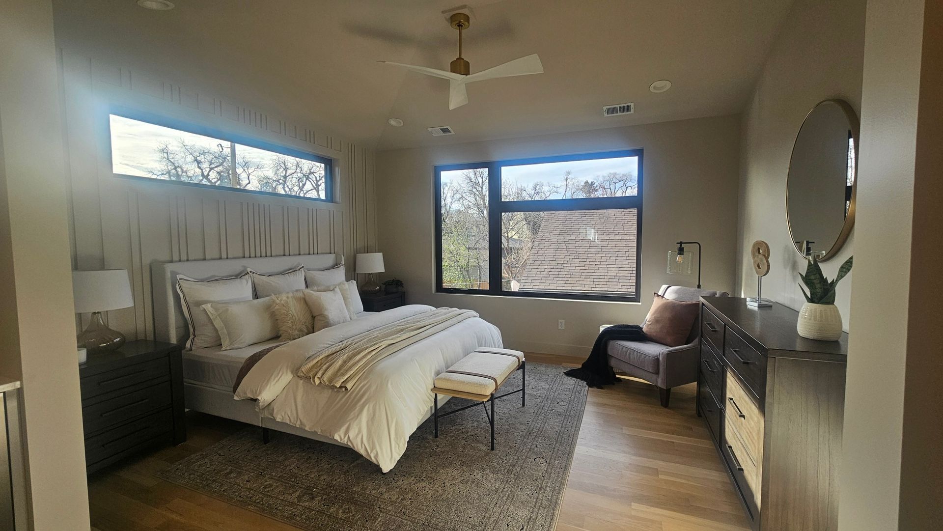 A modern, neutral-toned bedroom with a large bed, a ceiling fan, a gray area rug, a side chair, and a round wall mirror.