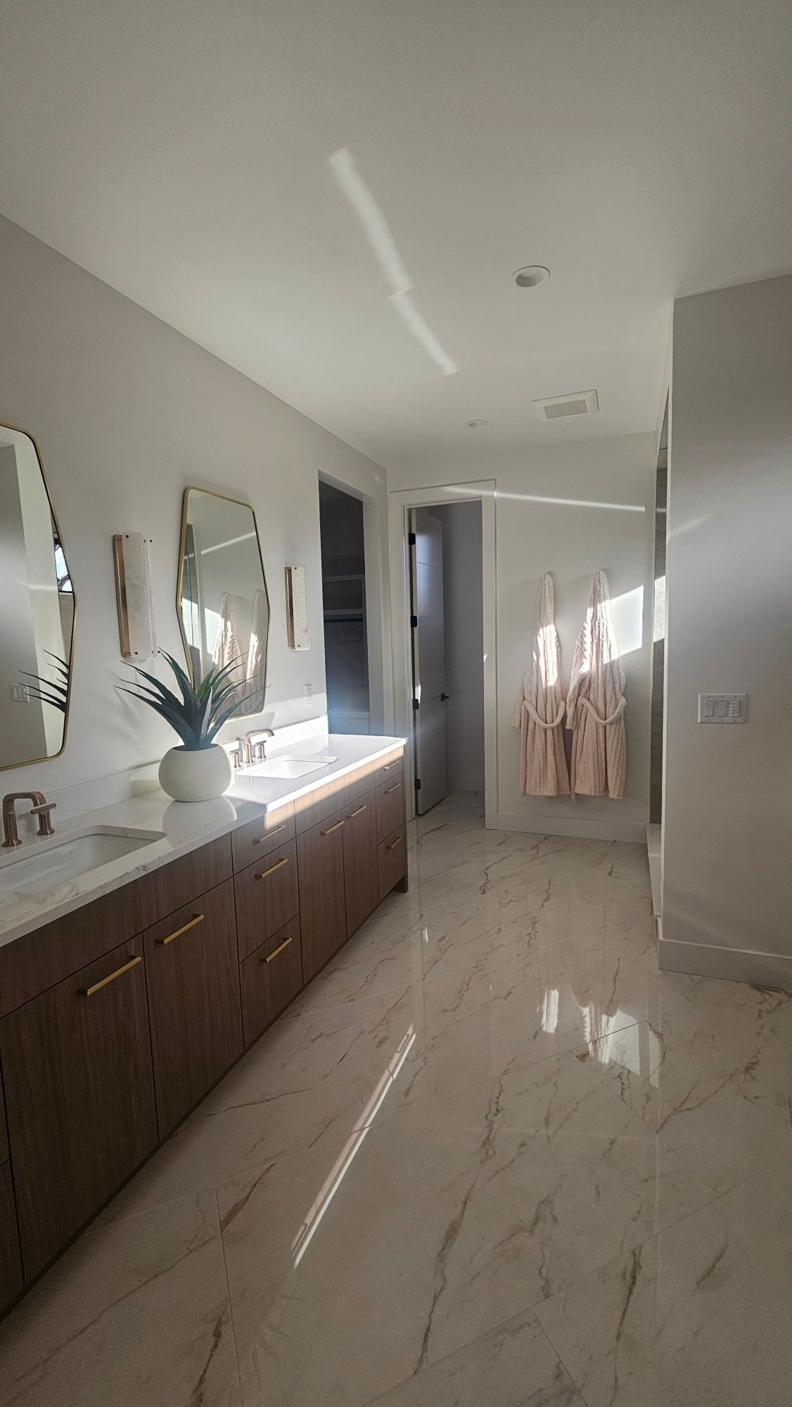 Modern bathroom with marble-look tile floors, dual wood vanities, light-colored walls, and robes hanging on the wall.