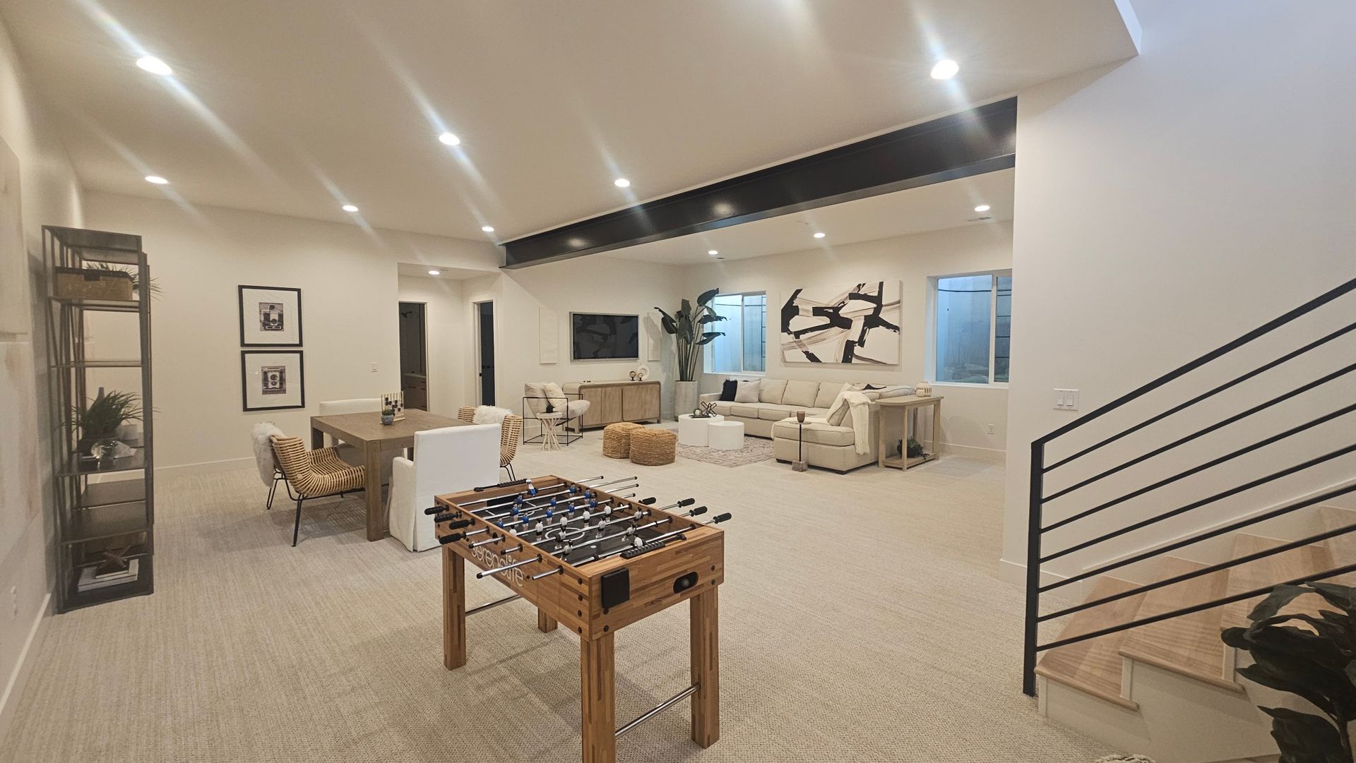 A spacious, neutral-toned basement featuring a foosball table, a dining area, and a comfortable living room lounge space.