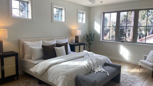 A well-lit, modern neutral-toned bedroom with a large bed, gray bench, and windows, featuring white and brown pillows.