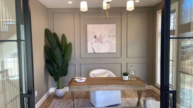 A home office with a wooden desk, white chair, tall potted plant, and abstract wall art against paneled, gray walls.