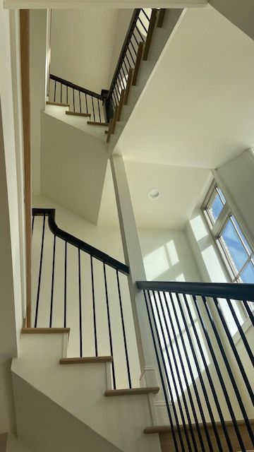 A modern staircase with white walls, wooden steps, and black metal railings, leading upward toward a bright window.