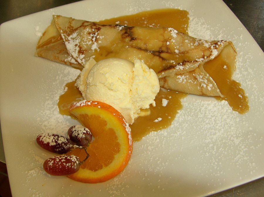 Crepes with ice cream, caramel sauce, orange slice, and sugared strawberries on a white plate.