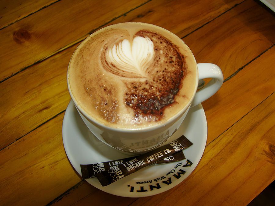 Cappuccino with heart-shaped latte art in white mug on saucer with sugar packets, on wooden table.