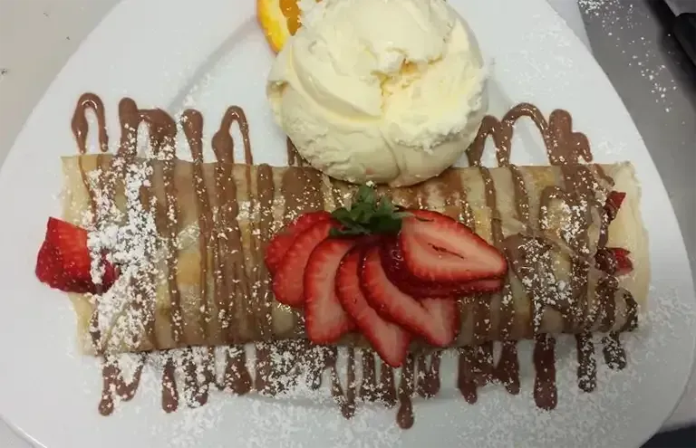 Crepe dessert with chocolate drizzle, strawberries, ice cream, and orange slice on a white plate.