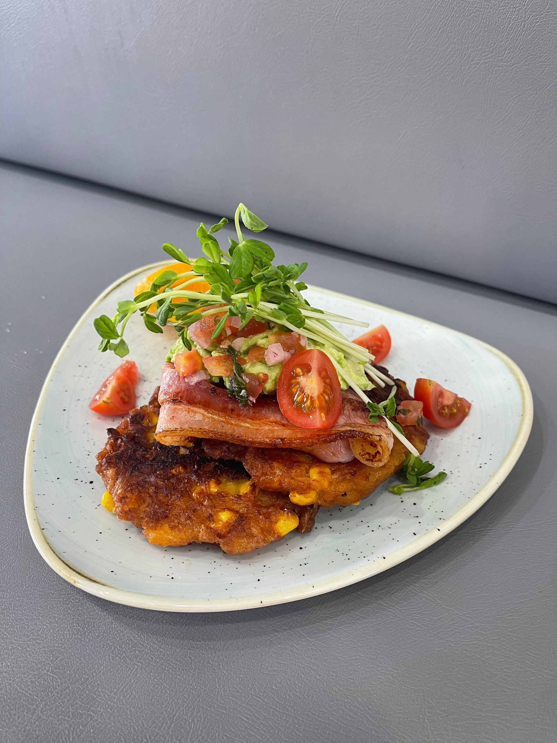 Corn fritters topped with bacon, avocado salsa, cherry tomatoes, and microgreens on a light blue plate.