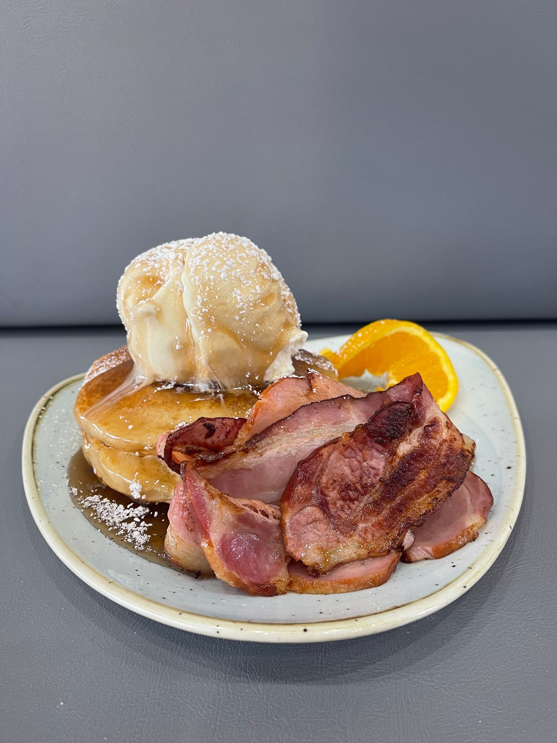 Pancakes topped with ice cream, bacon, and orange slice on a plate. Dusted with powdered sugar.