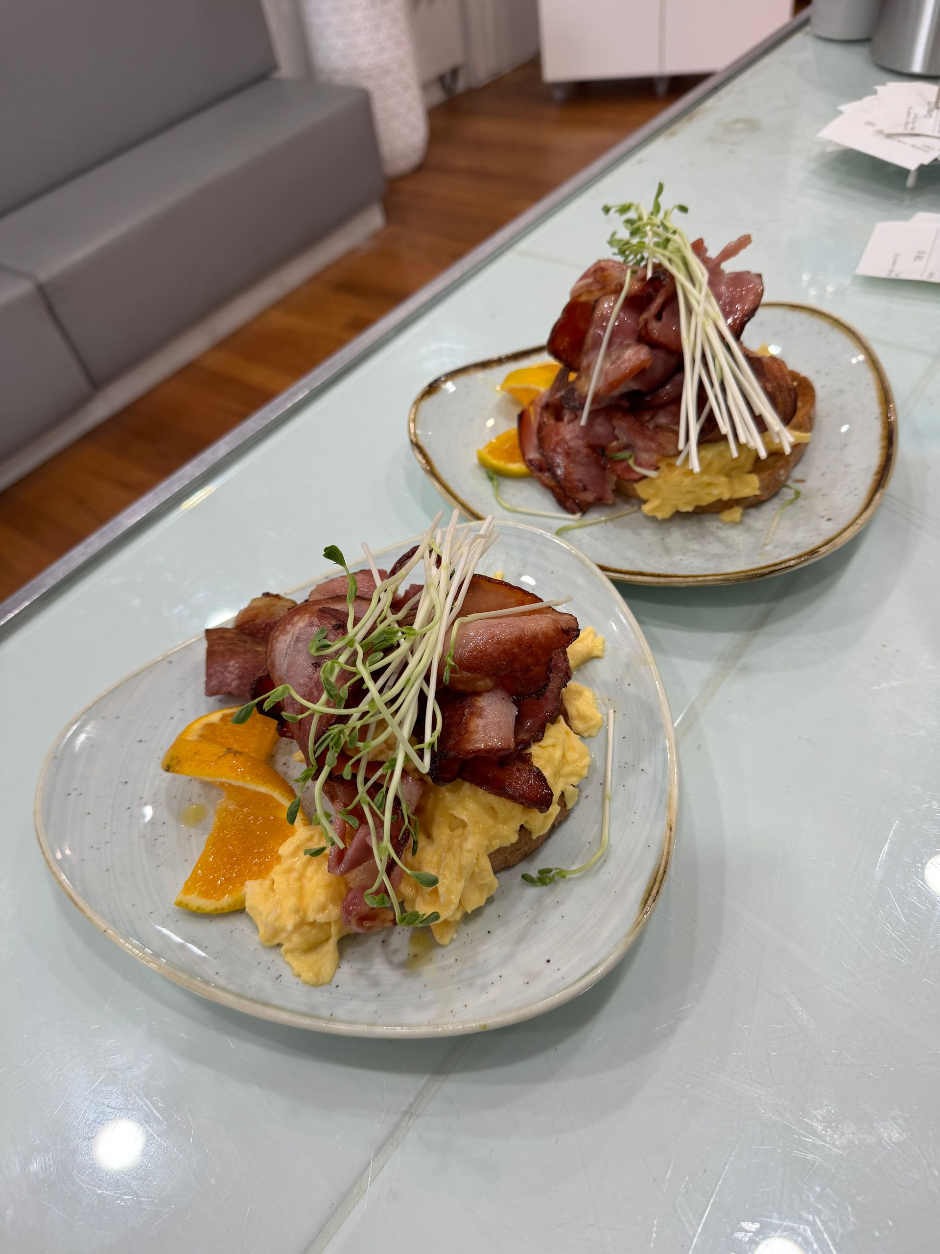 Two plates of scrambled eggs with bacon and orange slices, garnished with sprouts, on a white table.