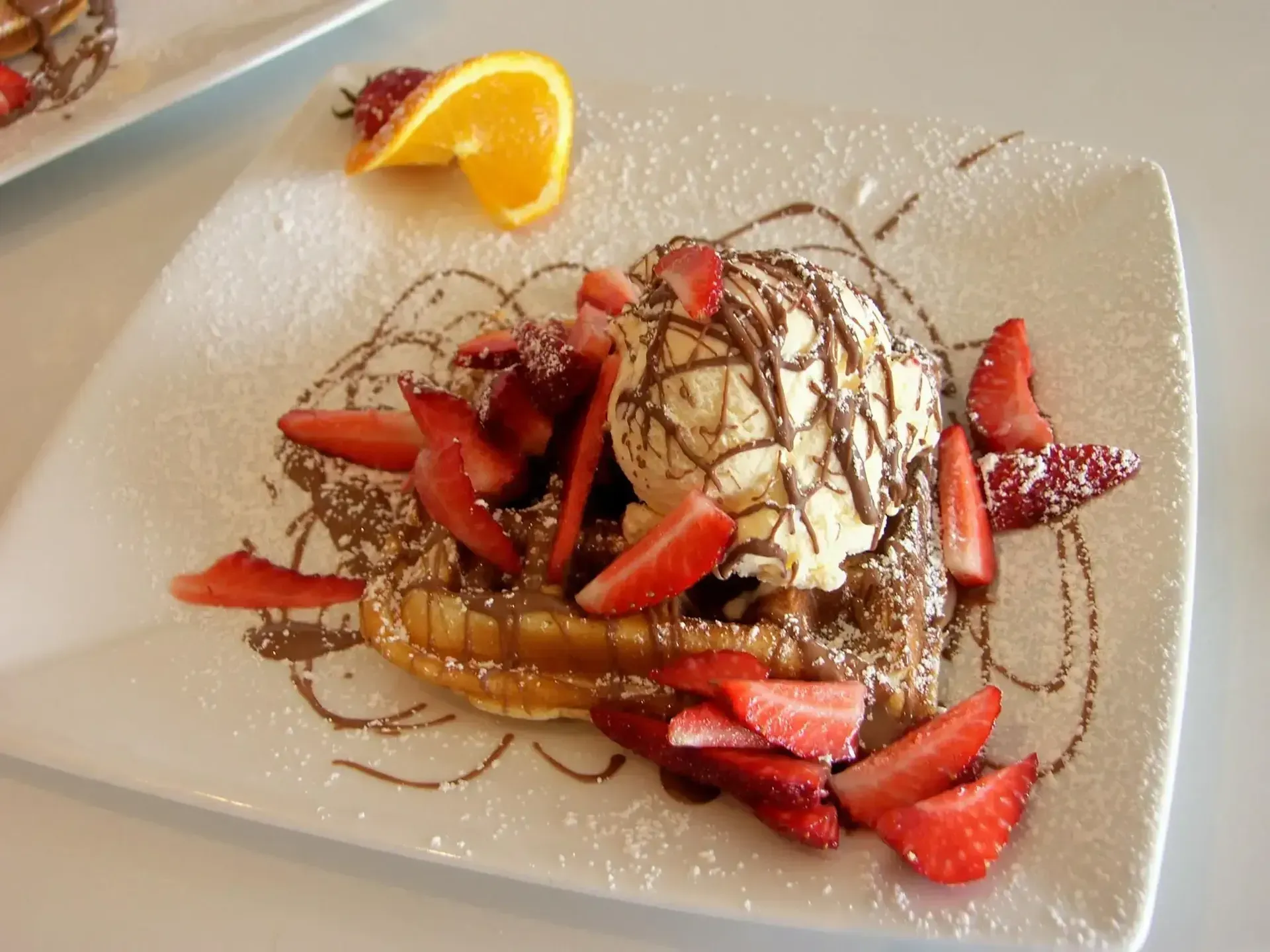 Waffle topped with ice cream, chocolate sauce, and strawberries on a white plate with orange slice garnish.