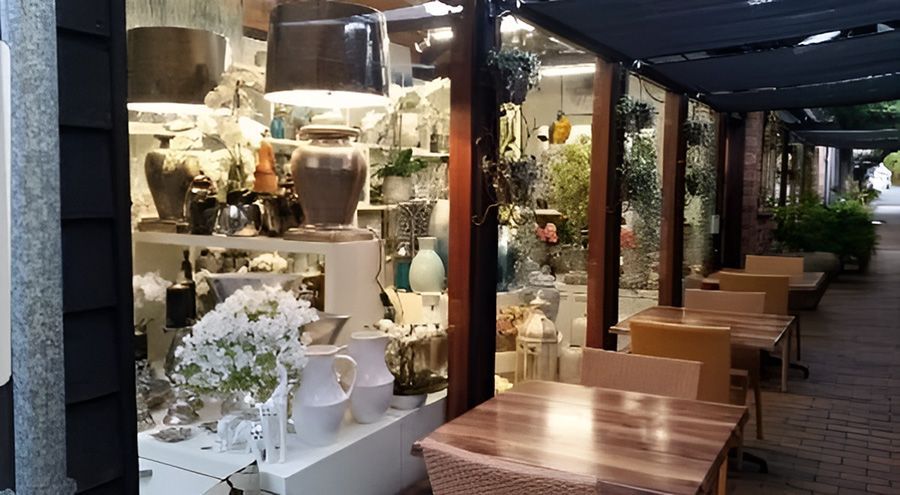 Shopfront display with pottery, flowers, and outdoor cafe seating under a wooden trellis.