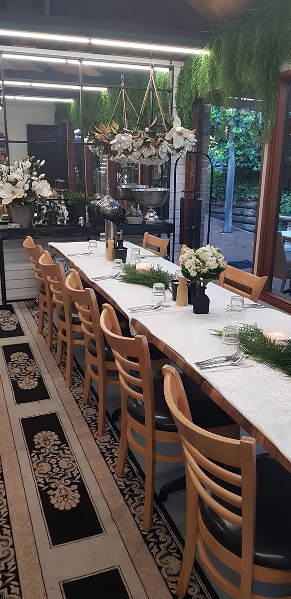 Long wooden table set for a formal event, with chairs along the sides. Decorations include flowers and greenery.