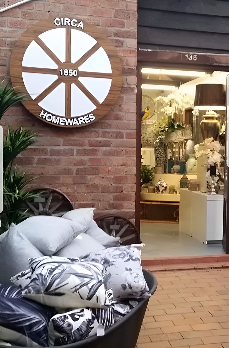 A Circa 1850 Homewares shop entrance with a wheel sign, brick wall, and display of pillows.