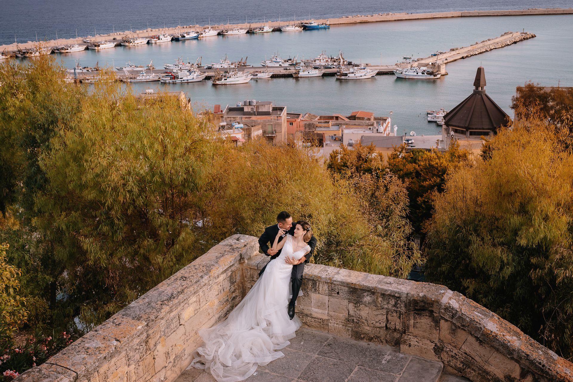 Una coppia di sposi è seduta su un muretto di pietra che domina un porto.