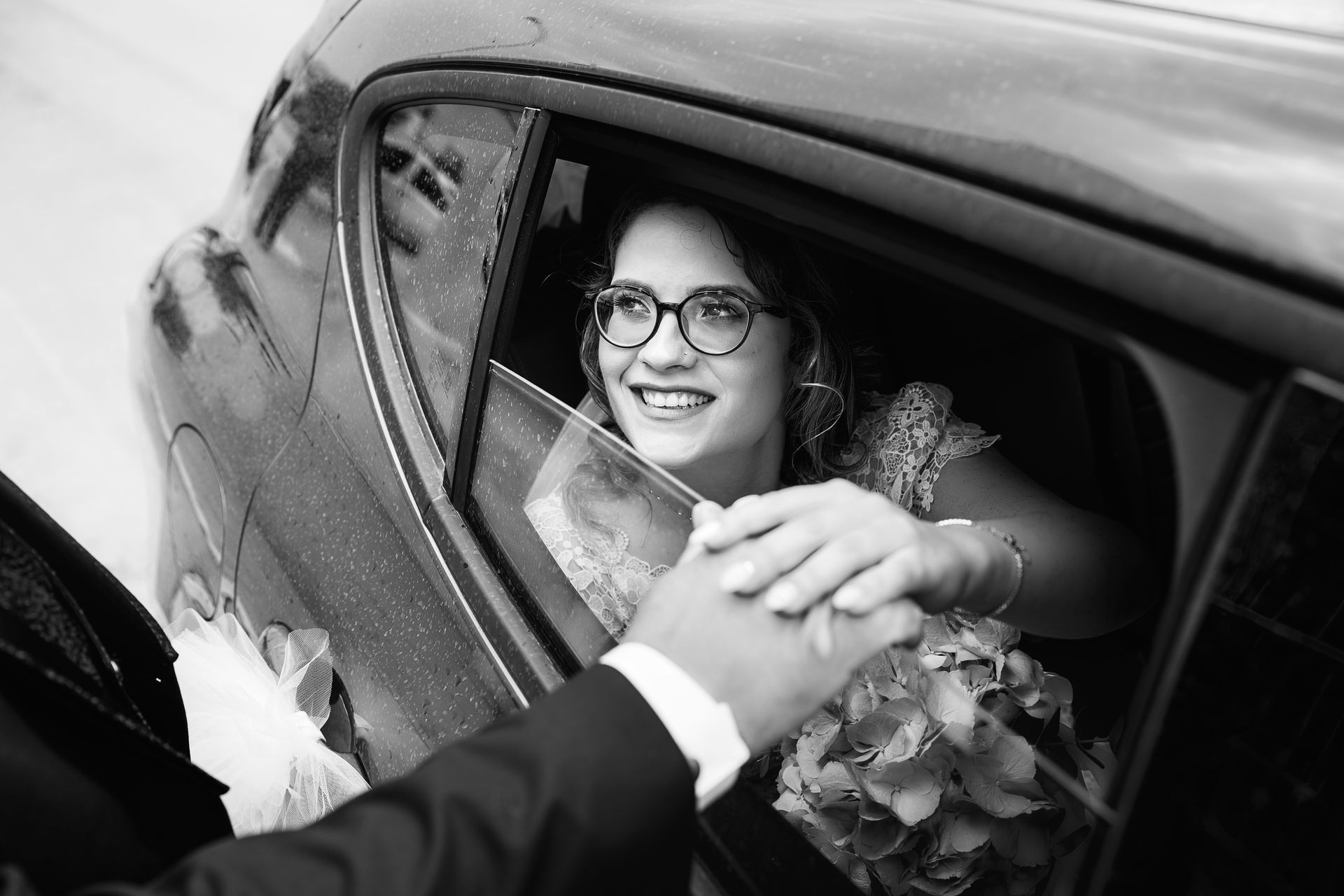 Una foto in bianco e nero di una sposa e uno sposo che si tengono per mano in macchina.