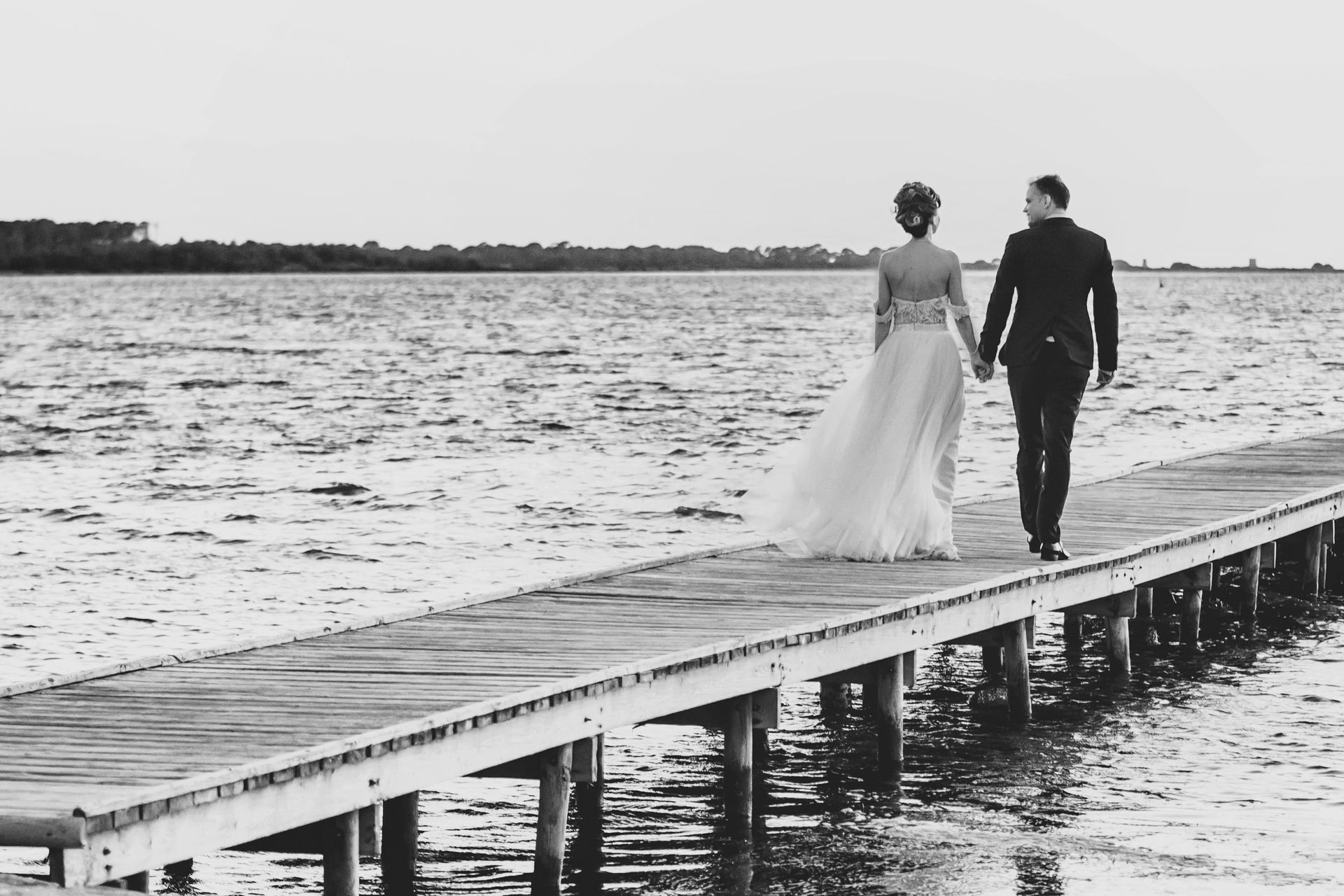 Una sposa e uno sposo camminano su un molo tenendosi per mano.