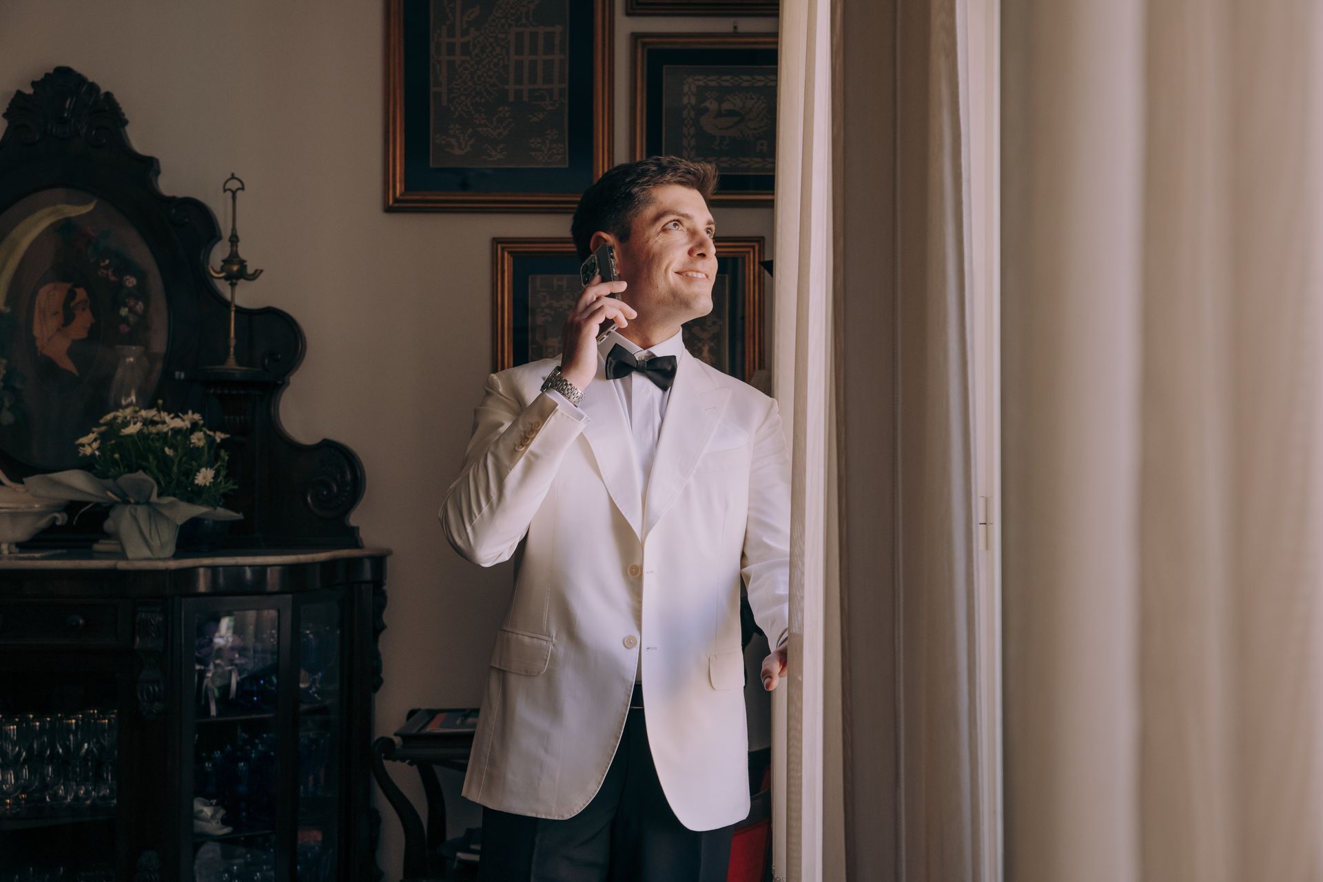 Un uomo in smoking è in piedi davanti a una finestra e parla al cellulare.