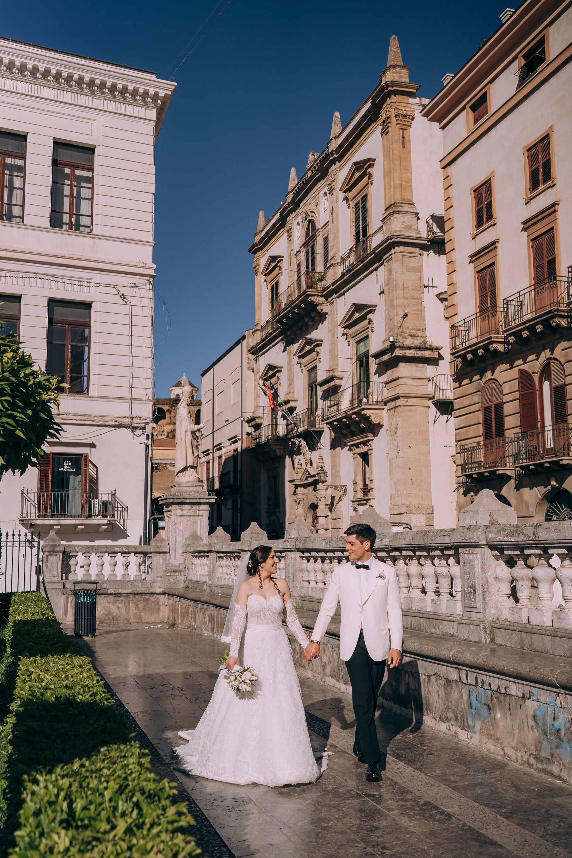 Una coppia di sposi cammina lungo un marciapiede tenendosi per mano davanti a un edificio.