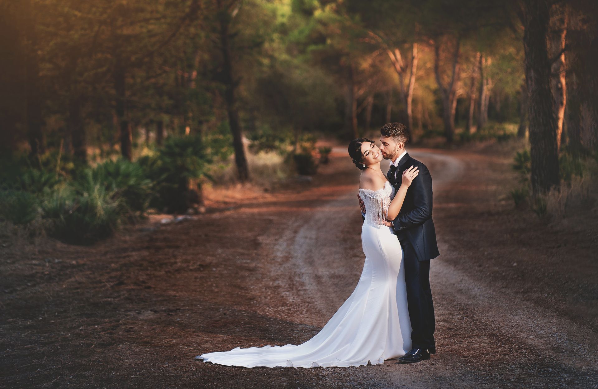 Una sposa e uno sposo si baciano su una strada sterrata nel bosco.