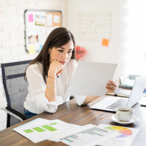 Woman at a desk reviewing papers, laptop, coffee cup, graphs, office setting, serious expression.