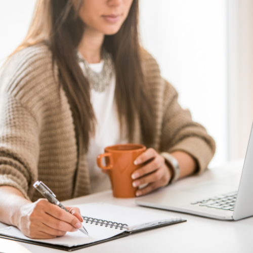 Woman writing in a notebook, holding mug, near laptop. Natural light. Brown cardigan, white top.