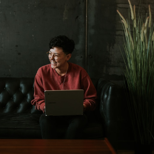 Person sitting on a black couch with a laptop, smiling. Dark red sweater and round glasses. Next to tall green plant.