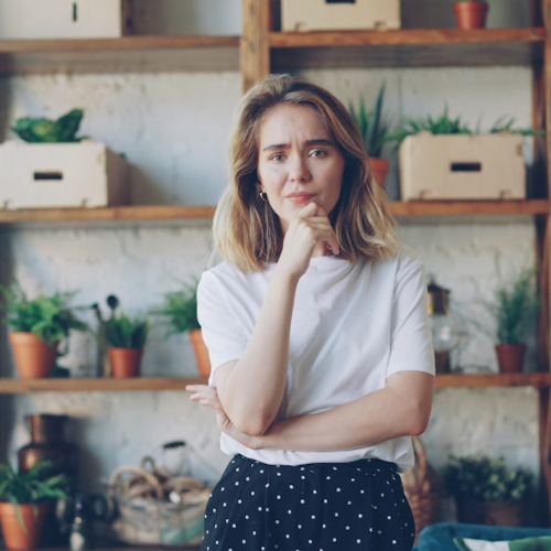 Woman in white shirt and polka-dot pants thoughtfully considers with a hand on her chin, plants and shelves in the background.