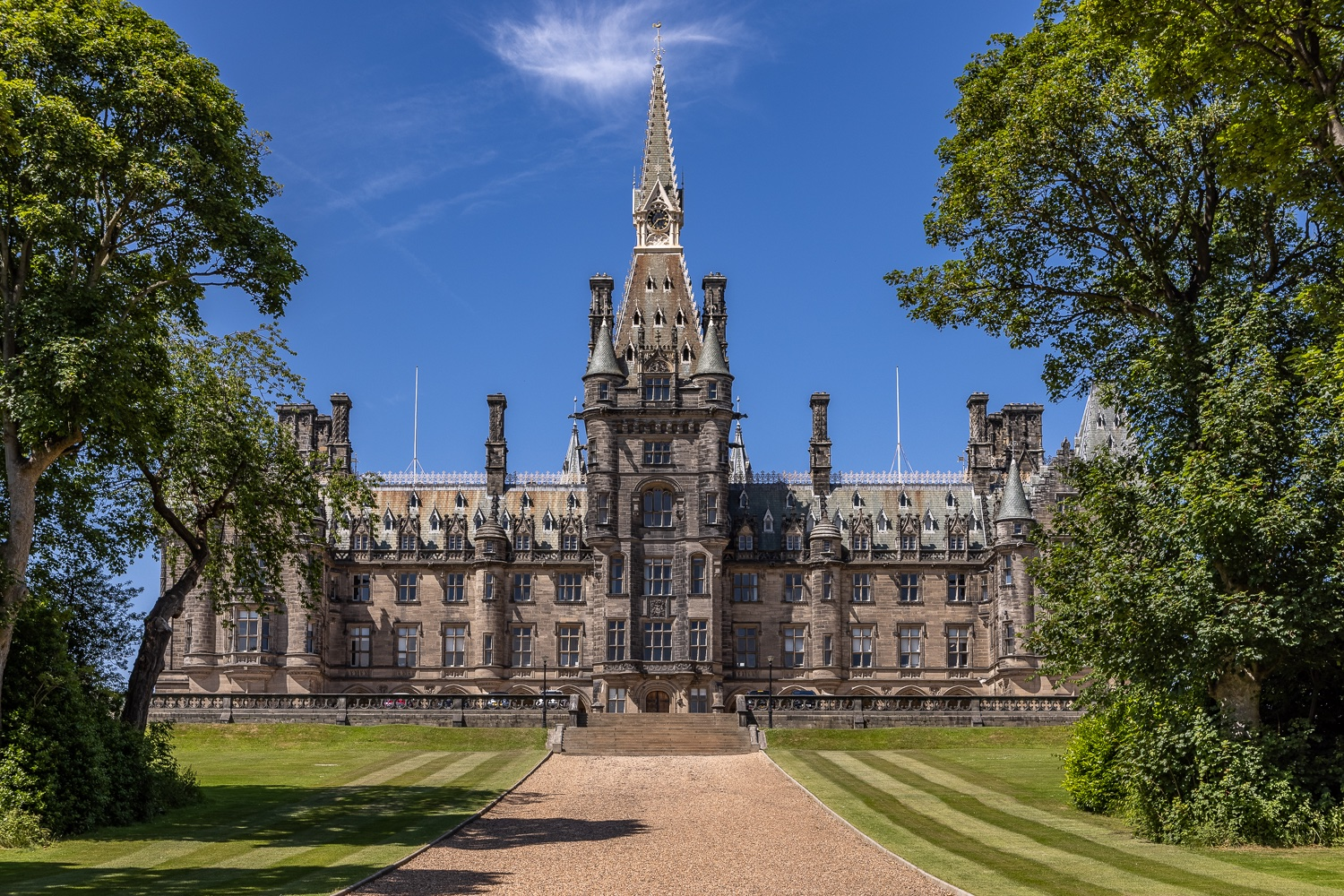 Gothic Revival mansion with central spire, framed by trees on a green lawn, under a blue sky.