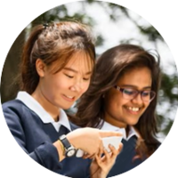 Two students looking at a phone. One points at the screen, both smile. Outdoors, wearing blue uniform tops.