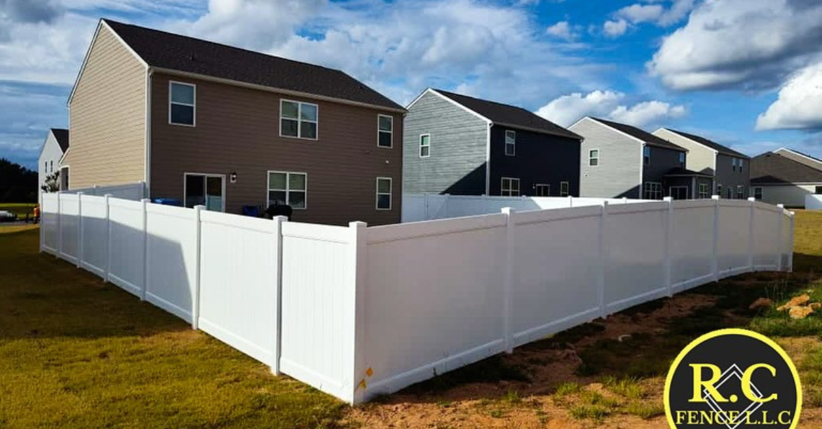 White vinyl fence surrounding a grassy yard with several houses in the background under a blue sky.