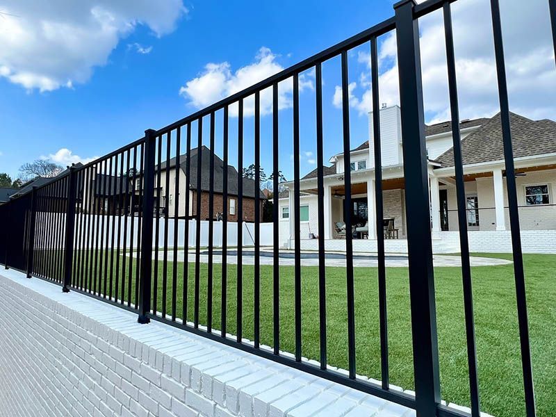 Black metal fence atop a white brick wall, enclosing a green lawn and a large house with white columns under a blue sky.