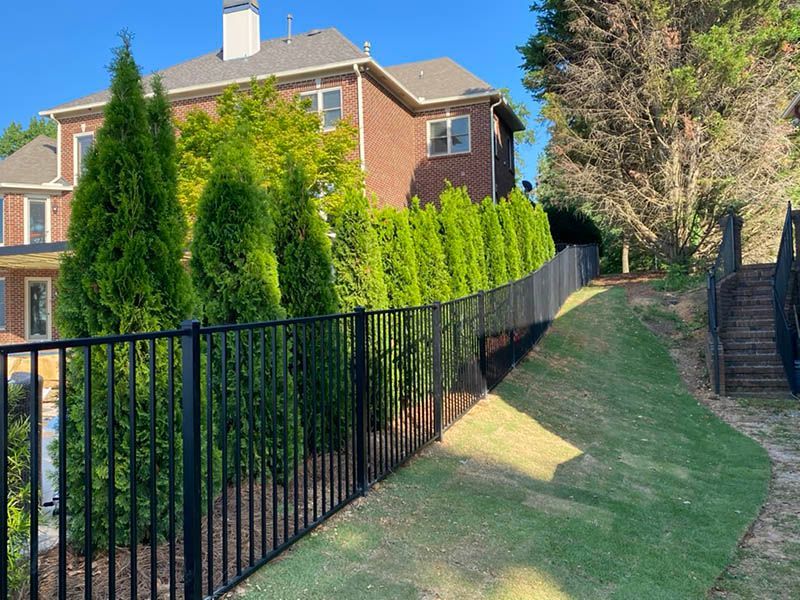 Black fence along a sloped lawn, with a brick house and evergreen trees in the background. Wooden stairs lead up a hill on the right.