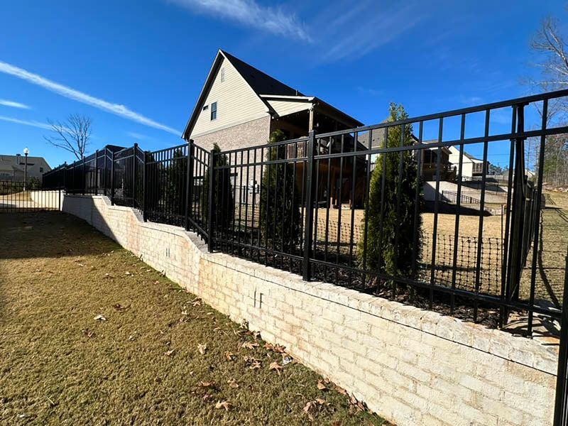 Black metal fence atop a beige retaining wall. Green trees are planted behind the fence, and a house with a dark roof is visible in the background.