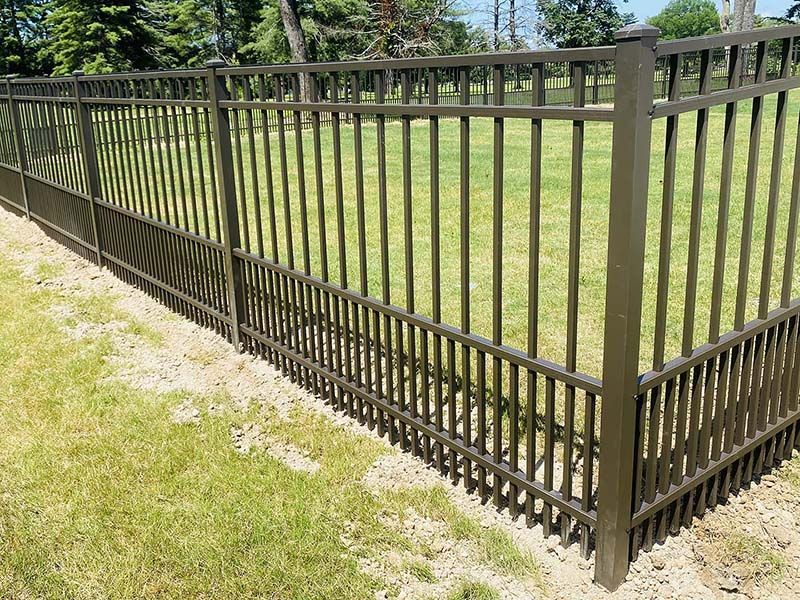 Brown metal fence on green grass, surrounding a yard with trees in the background.