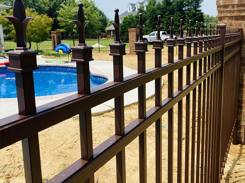 Brown metal fence surrounding a blue swimming pool on a sunny day. The fence has decorative spear-like finials.