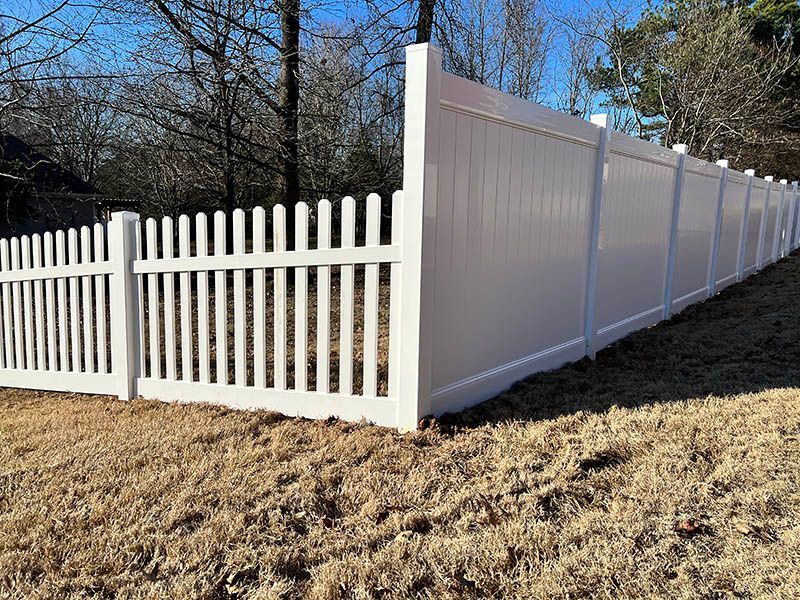 White picket fence with a solid panel section bordering a yard with brown grass and trees in the background.