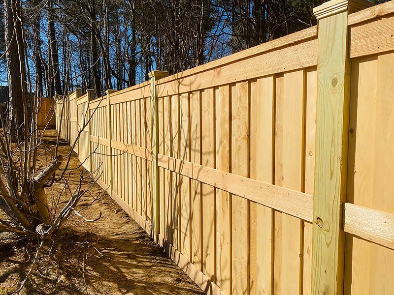 A wooden fence stands outdoors, with vertical planks and horizontal support beams. The fence is surrounded by trees and a clear sky.