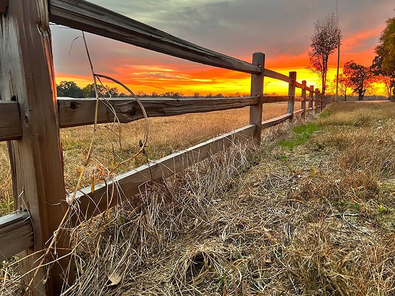 Wooden fence bordering a field, with a vibrant orange and yellow sunset in the background. Dry grass fills the foreground.