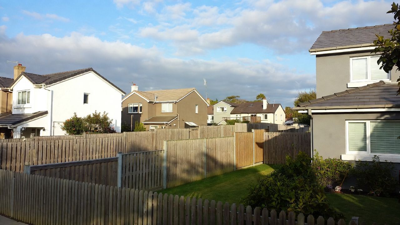 Suburban houses with gray and white exteriors, wooden fence, green lawn, cloudy sky.