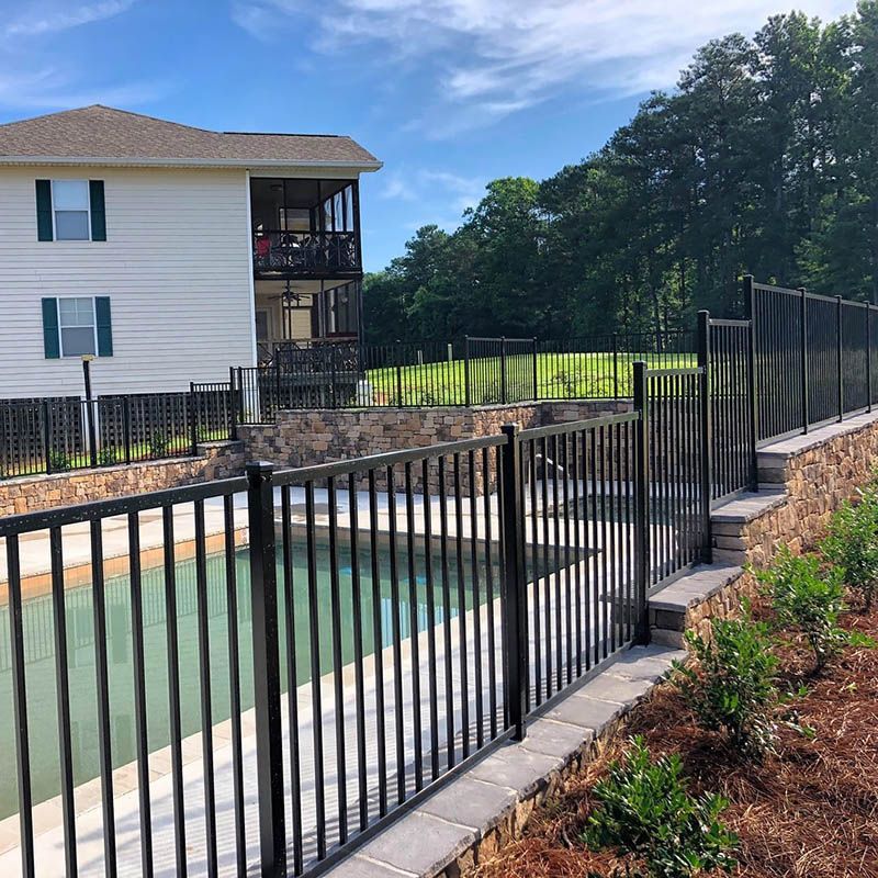 Black fence around a pool, with a multi-level building and green hillside in the background. Sunny day.