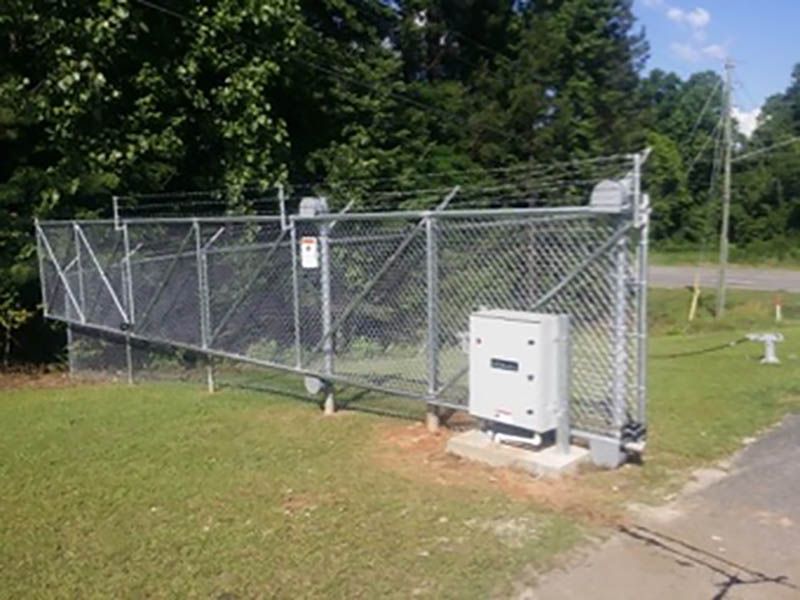 Chain-link fence with barbed wire at the top. A motorized gate and control box are visible. The gate is set in a grassy area next to a road.