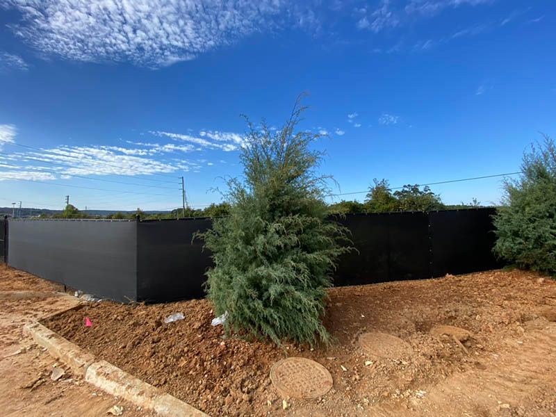 Black fence around a patch of dirt with a green bush under a bright blue sky.